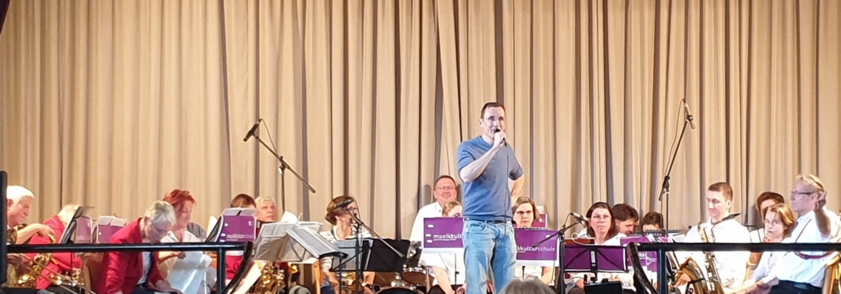 Das Bläserensemble der musikkulturschule Lauterbach und das Bläserensembel aus Niesig sitzen auf der Bühne des Posthotel Johannesberg. Christian Wiegand, Dirigent des Bläserensembles aus Niesig, moderiert.