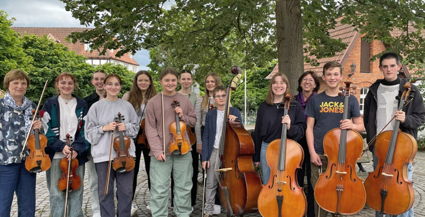 Eine Gruppe junger Musikerinnen und Musiker steht mit ihren Instrumenten unter einem Baum, sie halten ihre Instrumente, Geigen und Celli, in den Händen.